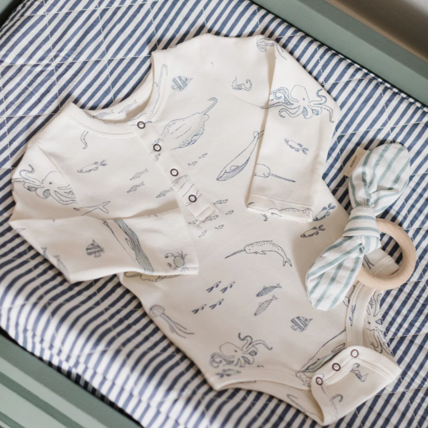 An ocean-themed baby bodysuit with long sleeves, printed with marine animals like octopuses, whales, and jellyfish. The onesie is placed on a striped blue-and-white background, accompanied by a wooden teething ring tied with a striped fabric bow