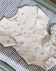 An ocean-themed baby bodysuit with long sleeves, printed with marine animals like octopuses, whales, and jellyfish. The onesie is placed on a striped blue-and-white background, accompanied by a wooden teething ring tied with a striped fabric bow