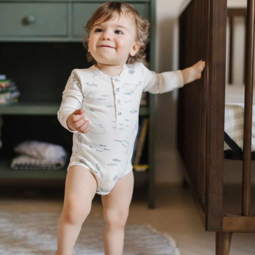 A smiling toddler wearing a long-sleeved ocean-themed bodysuit with illustrations of marine animals, standing confidently while holding onto the edge of a crib. A green shelf with neatly stacked books and folded fabric is visible in the background, adding a cozy nursery setting.