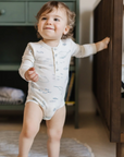 A smiling toddler wearing a long-sleeved ocean-themed bodysuit with illustrations of marine animals, standing confidently while holding onto the edge of a crib. A green shelf with neatly stacked books and folded fabric is visible in the background, adding a cozy nursery setting.