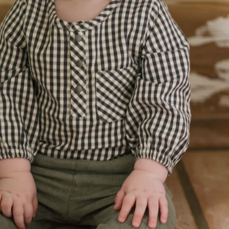 Close-up of a baby wearing a black-and-white gingham long-sleeved romper with a front button placket and chest pocket. The baby is seated, with their hands resting on green corduroy pants, adding texture to the outfit.