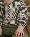 Close-up of a baby wearing a black-and-white gingham long-sleeved romper with a front button placket and chest pocket. The baby is seated, with their hands resting on green corduroy pants, adding texture to the outfit.