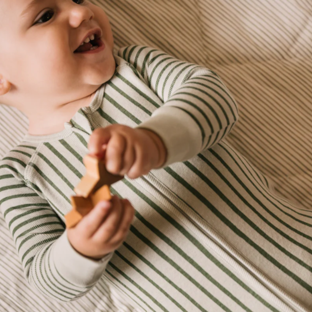 The image shows a smiling baby dressed in a striped footed onesie. The onesie has alternating sage green and light-colored vertical stripes, with solid light-colored cuffs at the wrists. The baby is holding a wooden toy and appears to be lying on a matching fabric with a similar striped pattern in the background, creating a cozy, coordinated look.