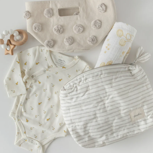 A flat lay of baby essentials, including a cream-colored baby onesie with small yellow animal prints, a wooden teether toy, a white muslin blanket with yellow patterns, and two pouches. One pouch is cream-colored with textured pom-pom accents, and the other is a soft gray-and-white striped pouch with a zipper, labeled "pehr." The arrangement presents a soft, neutral color palette, perfect for baby accessories and nursery organization.