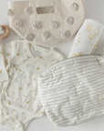 A flat lay of baby essentials, including a cream-colored baby onesie with small yellow animal prints, a wooden teether toy, a white muslin blanket with yellow patterns, and two pouches. One pouch is cream-colored with textured pom-pom accents, and the other is a soft gray-and-white striped pouch with a zipper, labeled "pehr." The arrangement presents a soft, neutral color palette, perfect for baby accessories and nursery organization.