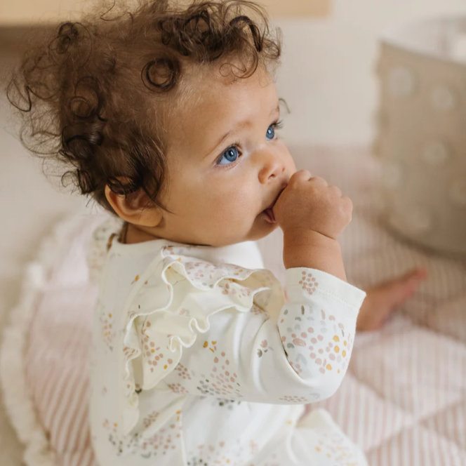 A baby with curly hair and bright blue eyes is sitting on a soft, striped quilted mat. The baby is wearing a cream-colored romper with pastel floral prints and delicate ruffle details along the shoulders. The baby has one hand in their mouth, adding a candid and sweet touch to the scene. The soft tones and cozy textures create a warm and soothing atmosphere.