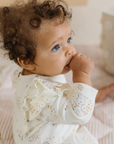 A baby with curly hair and bright blue eyes is sitting on a soft, striped quilted mat. The baby is wearing a cream-colored romper with pastel floral prints and delicate ruffle details along the shoulders. The baby has one hand in their mouth, adding a candid and sweet touch to the scene. The soft tones and cozy textures create a warm and soothing atmosphere.