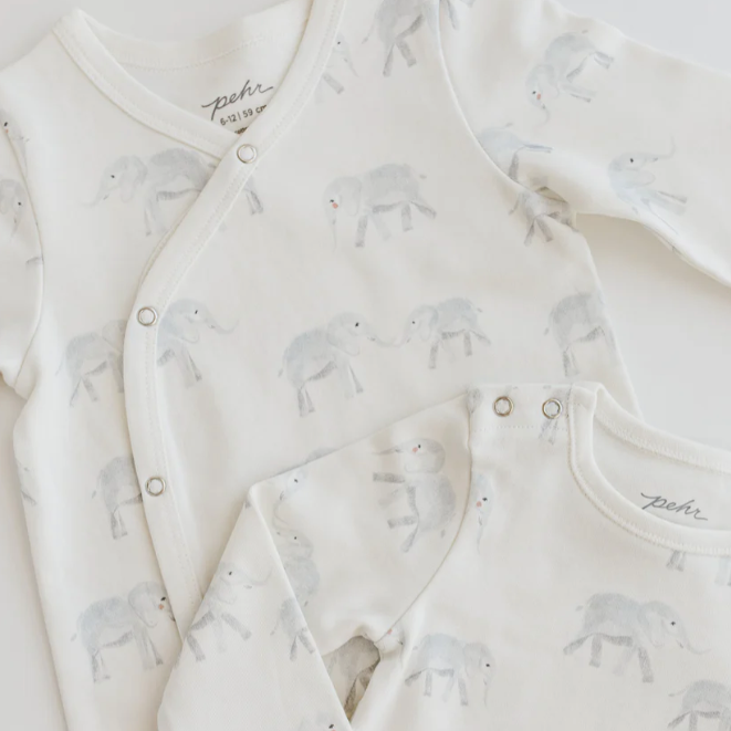 A close-up of two soft, cream-colored baby outfits featuring an all-over print of light gray elephants with small red accents on their cheeks. One outfit has a wrap-style design with snap closures along the front, and the other has a classic round neckline with snap buttons at the shoulder. The gentle colors and playful elephant pattern create a soothing and whimsical look for babywear.