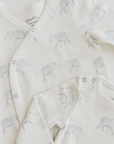 A close-up of two soft, cream-colored baby outfits featuring an all-over print of light gray elephants with small red accents on their cheeks. One outfit has a wrap-style design with snap closures along the front, and the other has a classic round neckline with snap buttons at the shoulder. The gentle colors and playful elephant pattern create a soothing and whimsical look for babywear.