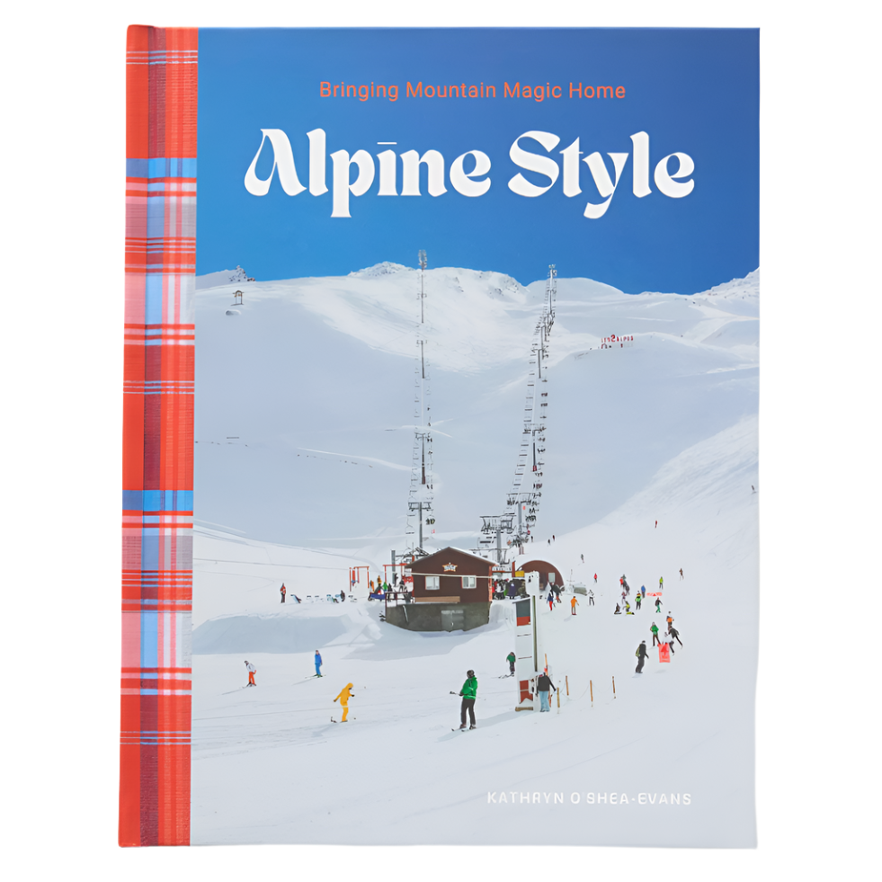 Front cover of the book "Alpine Style: Bringing Mountain Magic Home" by Kathryn O'Shea-Evans. The design features a snowy mountain scene with skiers, ski lifts, and a cozy alpine building. A bold plaid spine in red, blue, and white adds a touch of warmth to the design. The title suggests the book explores ways to incorporate alpine-inspired aesthetics into everyday living.