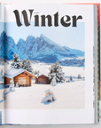 An interior page from "Alpine Style" featuring the title "Winter" in bold, stylized text. The image showcases a snow-covered landscape with rustic wooden cabins nestled among trees. Majestic mountains rise in the background, creating a serene alpine scene. This spread reflects the book’s focus on capturing the beauty and essence of winter in alpine settings.