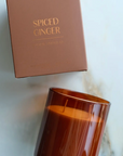 BROWN BOX LABELED: "BOTANICA SPICED GINGER HOLIDAY EDITION." THE CANDLE IS NEXT TO THE BOX AND IS ORANGE IN COLOR. THEY ARE LAID DOWN ON A MARBLE TABLE