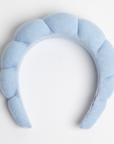 light blue padded terry cloth spa headband featuring a scalloped design along the top. The soft, plush texture gives it a comfortable appearance, making it suitable as a fashionable hair accessory. It is positioned against a plain white background.