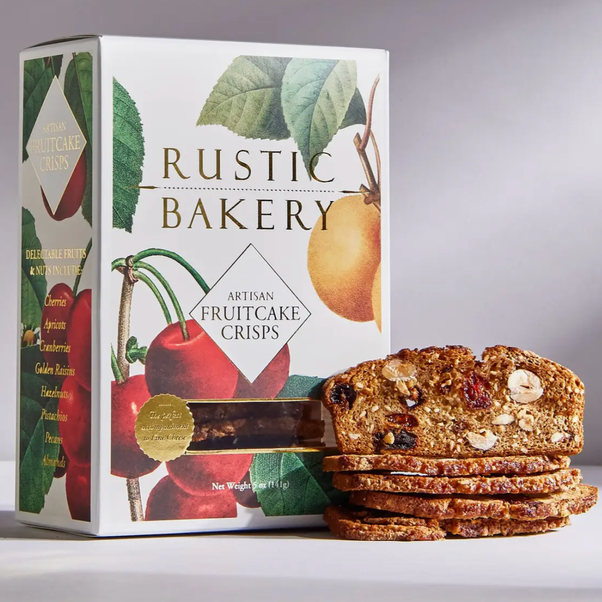 The image shows a box of Rustic Bakery Artisan Fruitcake Crisps alongside several slices of the crisps. The packaging features vibrant illustrations of cherries and another yellow fruit, possibly an apricot or peach, surrounded by green leaves. The product name, "Rustic Bakery Artisan Fruitcake Crisps," is printed prominently in elegant, gold-toned lettering. A transparent window on the box reveals the crisps inside, while a circular sticker emphasizes that it is "the perfect accompaniment to fine cheese." 