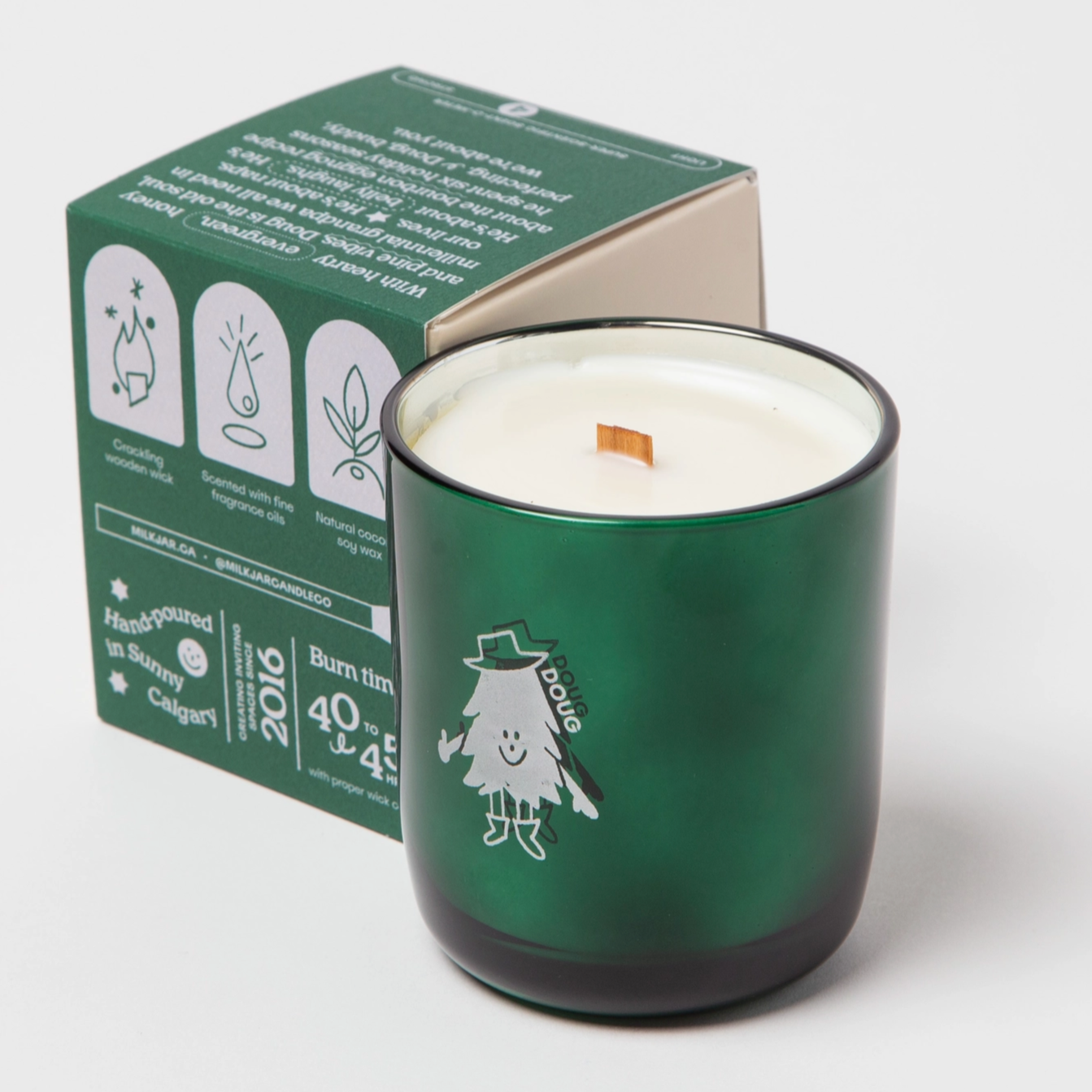 The image shows a green candle in a glass holder, with a box placed beside it. The candle features a small illustrated character, "Doug," wearing a hat and boots, adding a whimsical touch. The box has icons indicating features like a crackling wooden wick, scented coconut and soy wax, and hand-poured quality. It also mentions the candle's burn time (40 to 45 hours) and that it is handmade in Calgary. The text and branding emphasize the candle's artisanal nature and charity donation.

