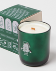 The image shows a green candle in a glass holder, with a box placed beside it. The candle features a small illustrated character, "Doug," wearing a hat and boots, adding a whimsical touch. The box has icons indicating features like a crackling wooden wick, scented coconut and soy wax, and hand-poured quality. It also mentions the candle's burn time (40 to 45 hours) and that it is handmade in Calgary. The text and branding emphasize the candle's artisanal nature and charity donation.

