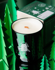 The image displays a green candle with a wooden wick, branded as "Doug" with an illustration of a small character wearing a hat and boots on the glass. The candle is surrounded by small, paper-cut green trees, evoking an evergreen forest. The box behind the candle matches its green color and design theme, highlighting the forest-inspired aesthetic of the product. The scene has a reflective surface, enhancing the candle's forest motif and creating a cozy, natural ambiance.