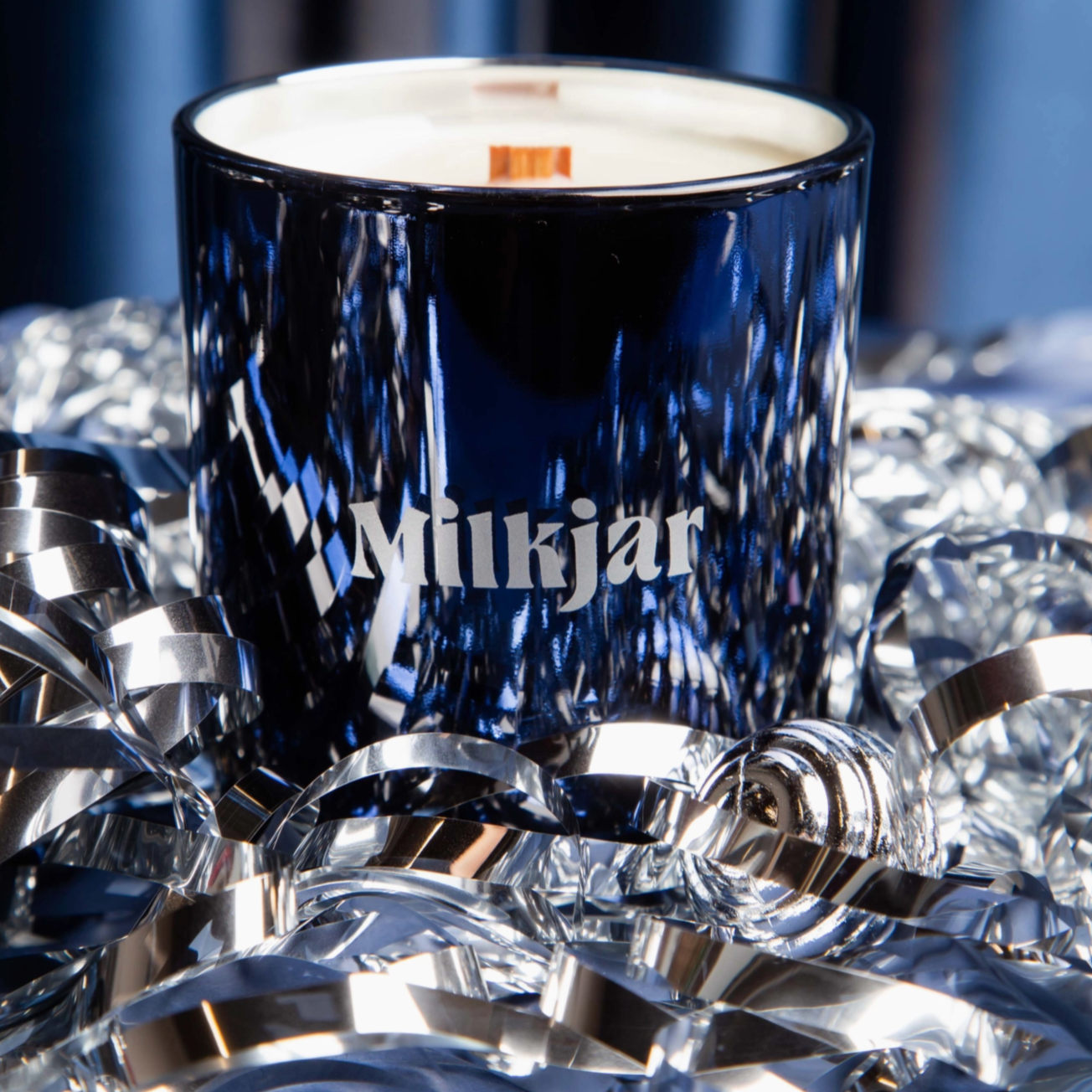 A blue glass candle with the brand name "Milkjar" printed on it sits among shiny silver ribbons against a blue background.
