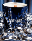 A blue glass candle with the brand name "Milkjar" printed on it sits among shiny silver ribbons against a blue background.
