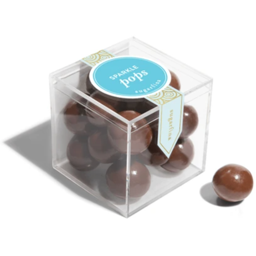 This image shows a clear plastic cube containing several glossy, round chocolate-coated candies labeled as "Sparkle Pops" by Sugarfina. A light blue circular label with gold detailing is affixed to the top, and a decorative banner runs down the side of the box. One candy is placed outside the box, showcasing its smooth, reflective chocolate coating.