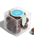 This image shows a clear plastic cube containing several glossy, round chocolate-coated candies labeled as "Sparkle Pops" by Sugarfina. A light blue circular label with gold detailing is affixed to the top, and a decorative banner runs down the side of the box. One candy is placed outside the box, showcasing its smooth, reflective chocolate coating.