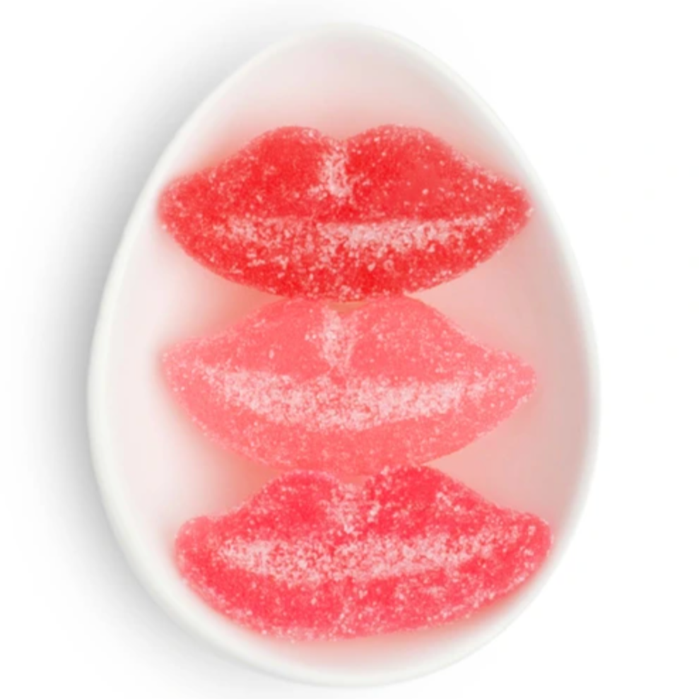 A close-up view of a small, oval-shaped white dish containing three lip-shaped, sugar-coated gummies arranged vertically. The gummies vary in shades of red and pink, giving a playful and vibrant look.