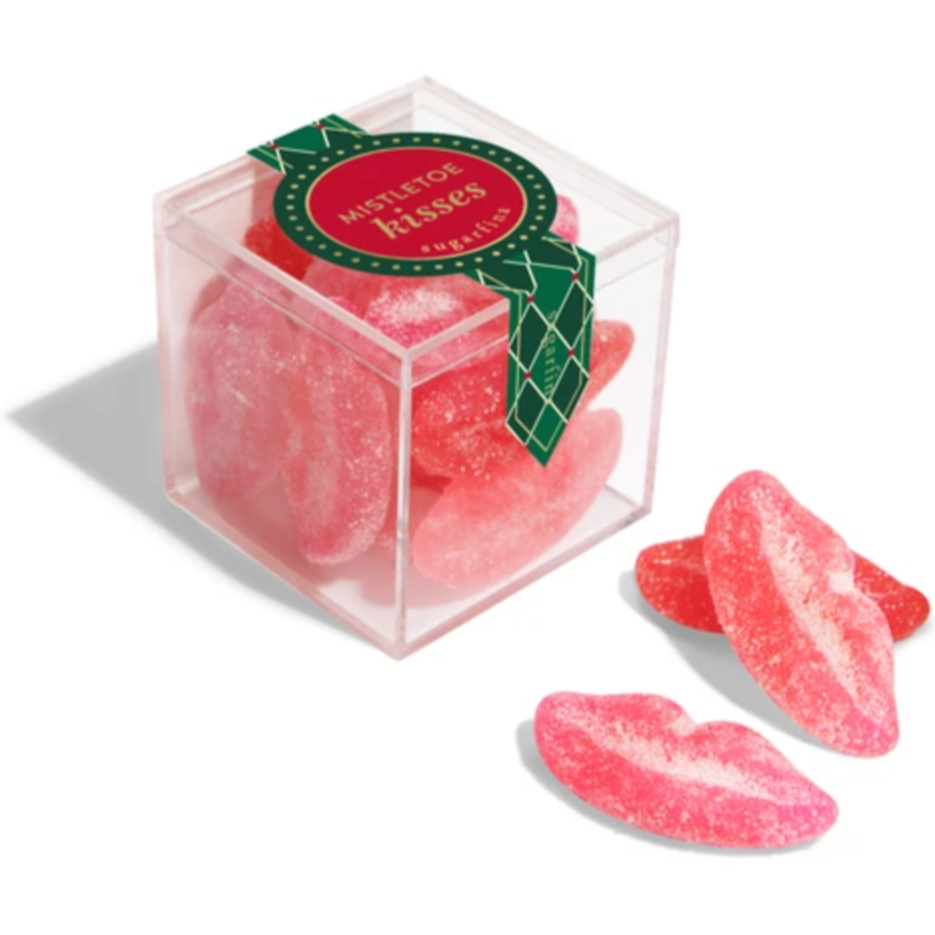 A transparent plastic cube with a festive label reading "Mistletoe Kisses." Inside the box are multiple pink and red lip-shaped, sugar-coated gummies. A few gummies are placed outside the box, showcasing their curved lip design and sugary texture.