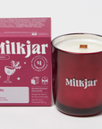 The image shows a maroon/red glass candle with a matching maroon/red box beside it. The box and candle both display the brand name "Milkjar." The box is labeled with the scent name "Holly," and lists fragrance notes: cranberry, clove and pine. The label mentions that the candle is made of coconut soy wax and that $1 from each candle is donated to charity. The candle has a wooden wick visible at the center.