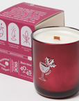 The image shows a maroon/red glass candle with a matching maroon/red box beside it. The box and candle both display the brand name "Milkjar." The box is labeled with the scent name "Holly," and lists fragrance notes: cranberry, clove and pine. The label mentions that the candle is made of coconut soy wax and that $1 from each candle is donated to charity. The candle has a wooden wick visible at the center. The candle has a design of a cup with a smiley face that says Holly on it. 