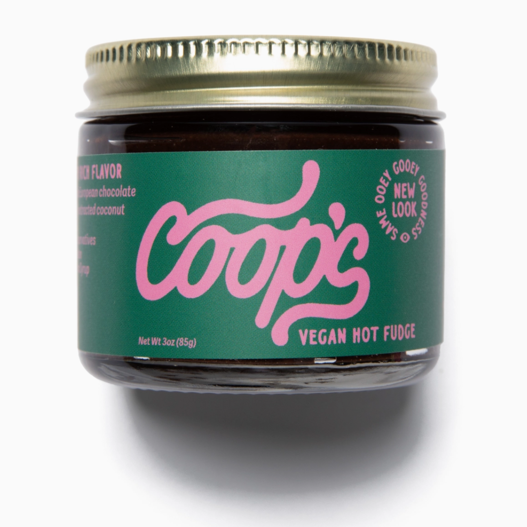 The image shows a jar of "Coop's Vegan Hot Fudge" with a pink and green label. The brand name "Coop's" is written prominently in a stylized pink font on a green background. To the right, there's a circular text that says, "SAME OOEY GOOEY GOODNESS" and "NEW LOOK" around it. The label also mentions "VEGAN HOT FUDGE" and highlights the absence of preservatives and corn syrup. The net weight is indicated as 3 oz (85 g).