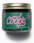 The image shows a jar of "Coop's Vegan Hot Fudge" with a pink and green label. The brand name "Coop's" is written prominently in a stylized pink font on a green background. To the right, there's a circular text that says, "SAME OOEY GOOEY GOODNESS" and "NEW LOOK" around it. The label also mentions "VEGAN HOT FUDGE" and highlights the absence of preservatives and corn syrup. The net weight is indicated as 3 oz (85 g).