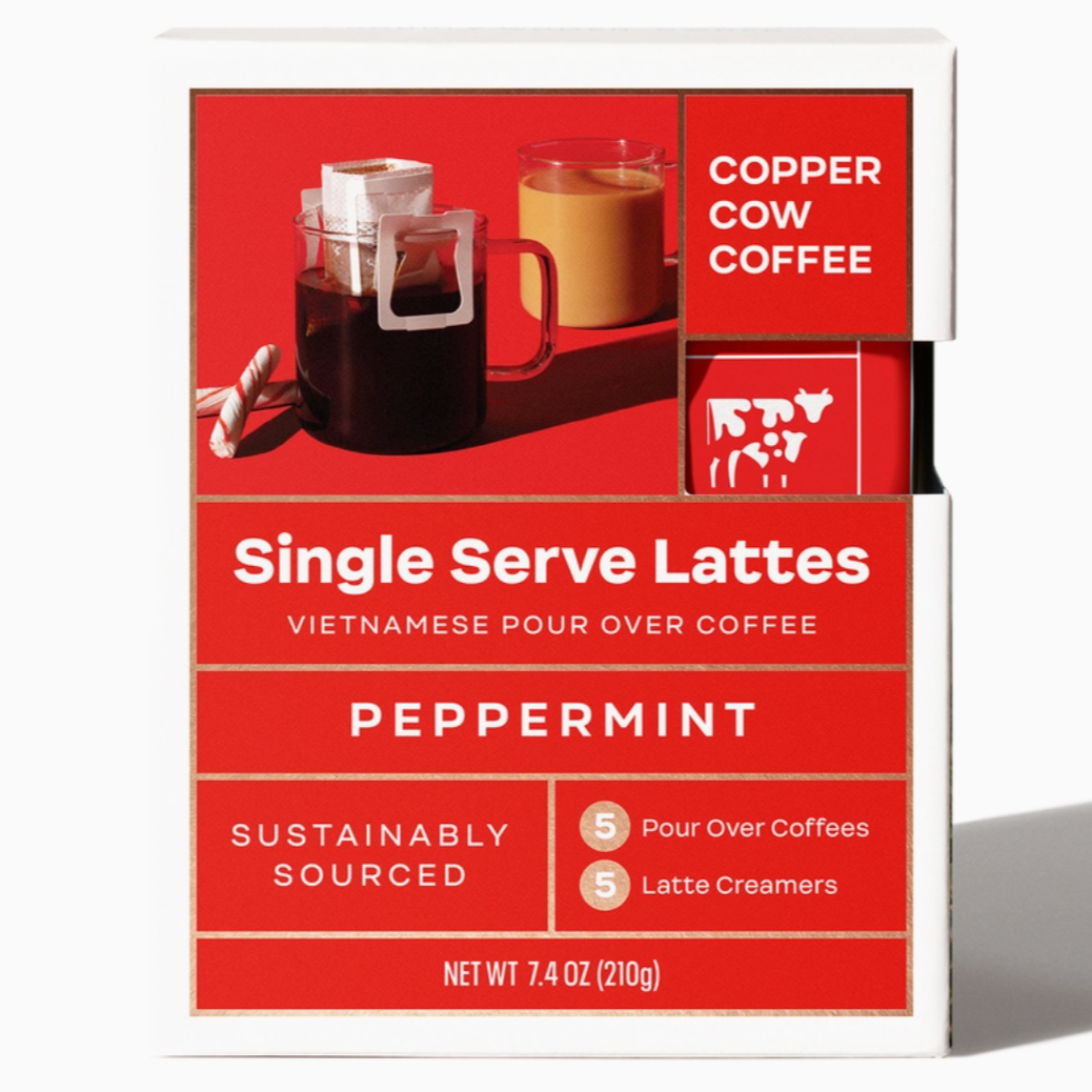 a box of Copper Cow Coffee Single Serve Lattes in the Peppermint flavor. The packaging is primarily red and white, featuring an image of two glass mugs – one with black coffee and a pour-over filter, and the other with a creamy latte. Two candy canes are next to the black coffee mug, adding a festive feel. The text on the box reads: "Single Serve Lattes, Vietnamese Pour Over Coffee, Peppermint, Sustainably Sourced," and indicates that it includes "5 Pour Over Coffees" and "5 Latte Creamers." 