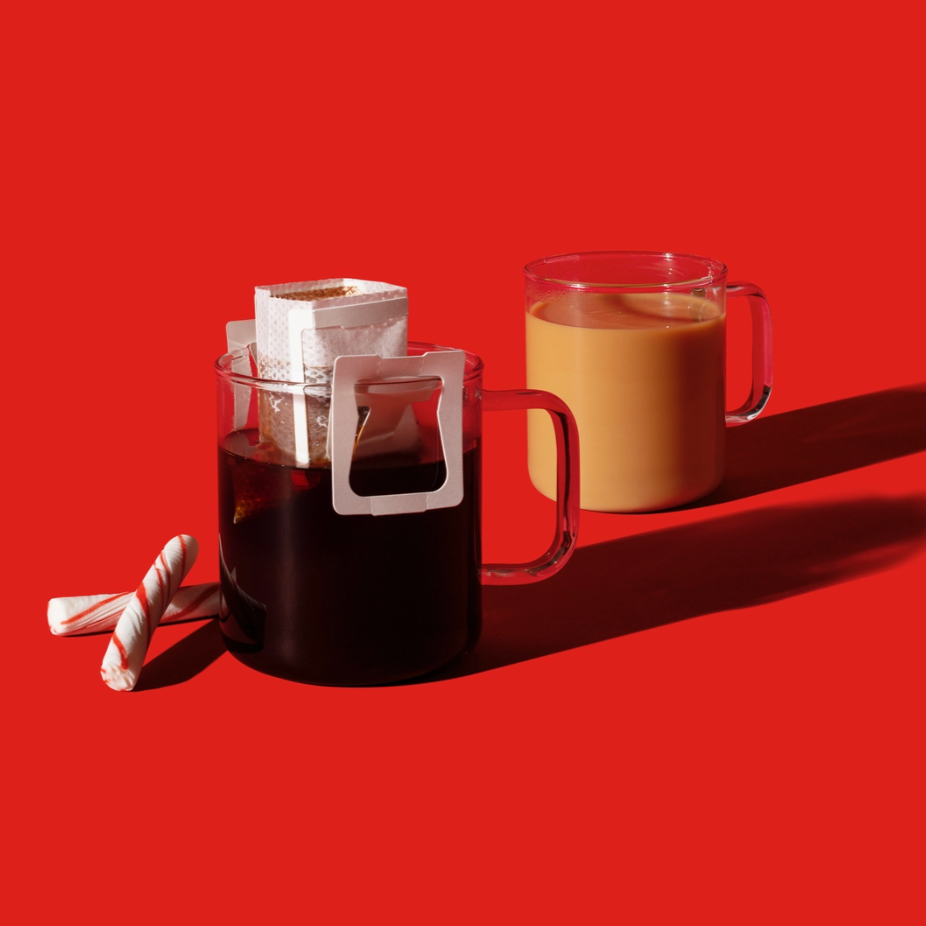  Two clear glass mugs on a bright red background. One mug contains black coffee with a white pour-over filter attached, while the other has a creamy brown coffee. Two candy canes are placed beside the black coffee mug, adding a festive touch.