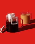  Two clear glass mugs on a bright red background. One mug contains black coffee with a white pour-over filter attached, while the other has a creamy brown coffee. Two candy canes are placed beside the black coffee mug, adding a festive touch.