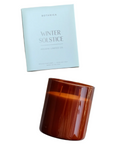 The image shows a candle product from the brand "Botanica" with the label "Winter Solstice" and "Holiday Limited Ed" on the packaging. The packaging is a simple, light-colored box with minimalist text, conveying a calm and festive feel. The candle is in a warm amber-colored glass container and is placed next to the box on white background. This setup enhances the candle's luxurious and cozy aesthetic, creating an inviting winter ambiance.
