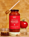 A jar of SAUZ Hot Honey Marinara sauce labeled as 'Sweet & Spicy Tomato Sauce,' positioned on a wooden surface. A honey dipper drips honey onto the top of the jar. To the left, there is a small glass jar filled with red chili flakes and oil. To the right of the jar, a ripe tomato, a piece of garlic, and a slice of onion are arranged, all against a warm, wood-grain background