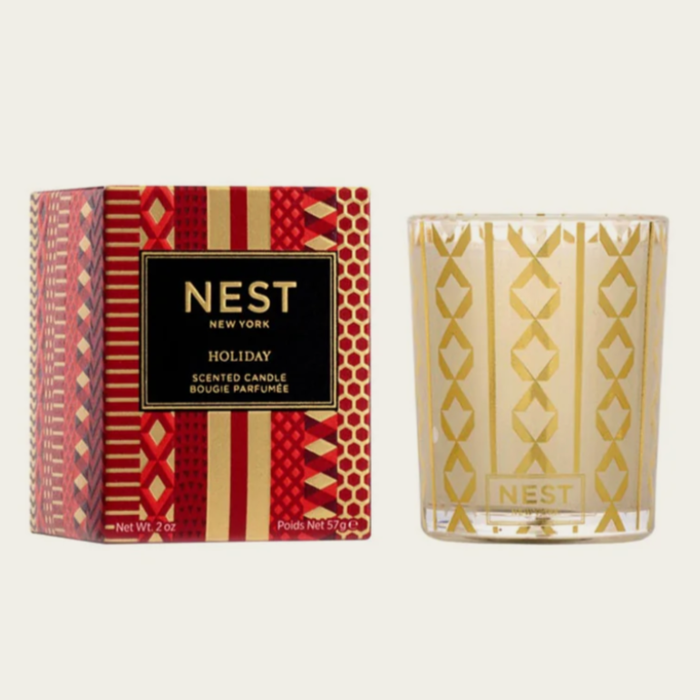 This image shows a holiday-themed scented candle from the brand "Nest New York." The candle is in a clear glass holder with a decorative gold pattern, and it comes in a red and gold box with festive designs. The box displays the brand name "Nest" and indicates it is a "Holiday" scented candle, with the description "Scented Candle" and a weight of 57g or 2 oz.


