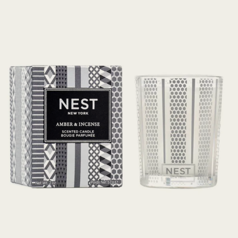 Nest New York scented candle in the fragrance "Amber & Incense." The packaging is predominantly gray and white with a geometric, textured pattern. The candle jar is transparent with a matching geometric pattern that complements the box design, and it has the Nest logo on the front.






