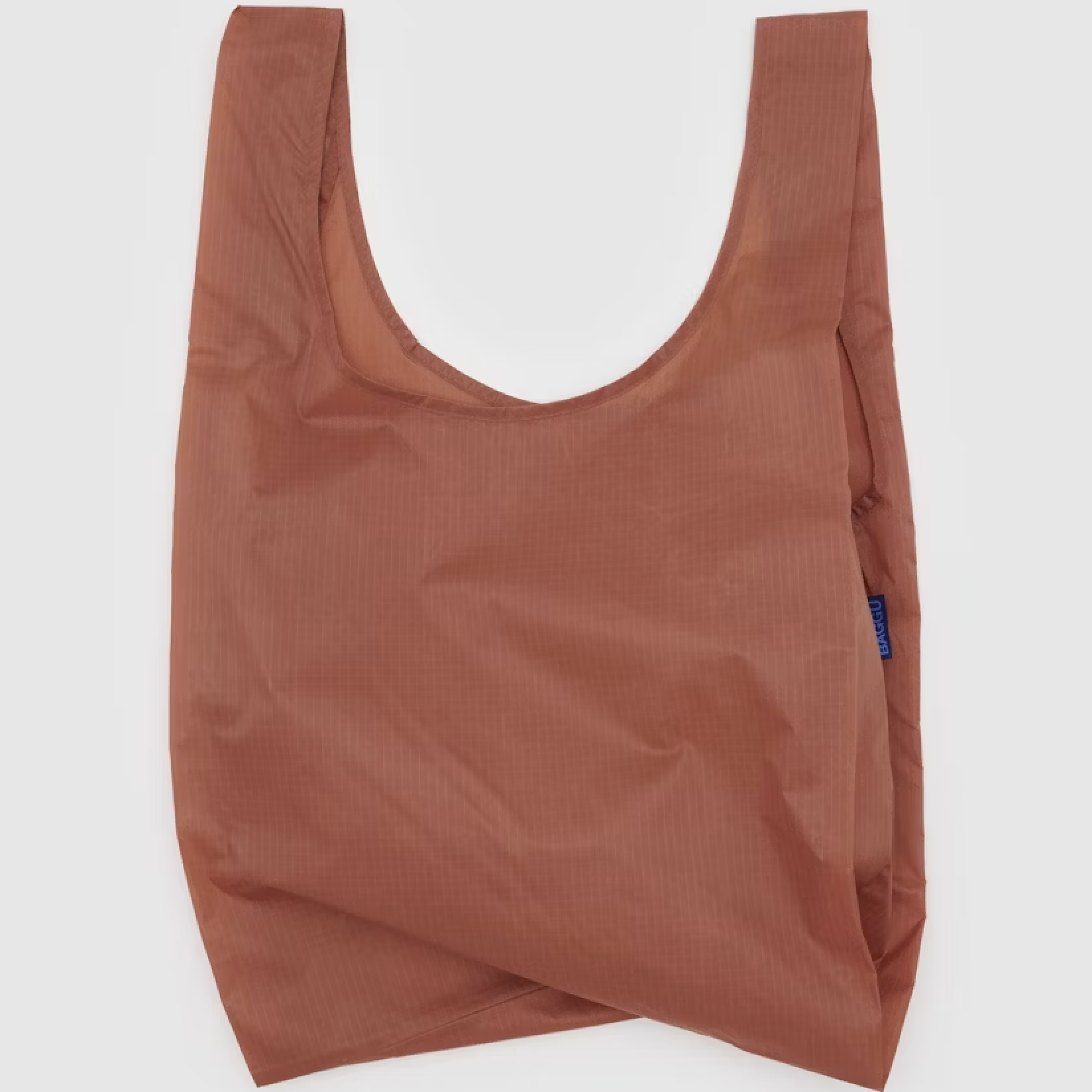 This image shows a terracotta brown reusable shopping bag with two handles, likely made from a lightweight, durable fabric. The design is simple and unstructured, and a small blue label with "BAGGU" written on it is visible on the side.







