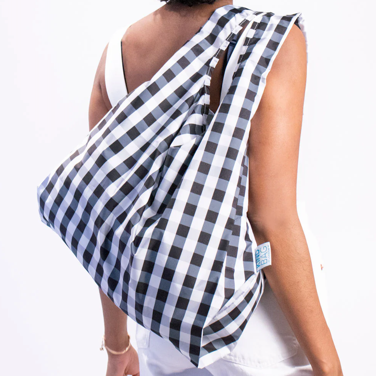 A person with curly black hair is wearing a large, checkered reusable bag over their shoulder. The bag has a black-and-white gingham pattern and is labeled with a small tag that reads "BIG BAG." The person is dressed in white clothing, contrasting with the bag’s dark pattern.


