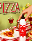 bottle of "Brightland Pizza Oil" on a red and white checkered tablecloth. A hand with red-painted nails is reaching for the bottle. In the background, there is a neon "PIZZA" sign on a green wall. The table is set with glasses of red wine, a slice of pepperoni pizza on a plate, a pizza cutter, and a bowl of salad. The bottle has a red label with white text, reading "Brightland Pizza Oil" and described as "Jalapeño & Herb Flavored Olive Oil," with a volume of "12 FL OZ (355 mL)."