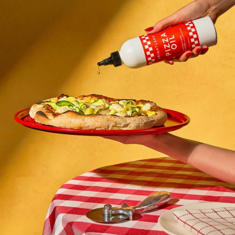 hand pouring "Brightland Pizza Oil" onto a freshly made pizza topped with zucchini and cheese, displayed on a red plate. The background has a warm yellow color, adding to the cozy and inviting atmosphere. The table beneath has a red and white checkered tablecloth, with a pizza cutter and a plate in view. The bottle features a red label with white text and checkered borders, describing the oil as "Brightland Pizza Oil," "Jalapeño & Herb Flavored Olive Oil," with a volume of "12 FL OZ (355 mL)