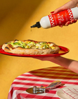 hand pouring "Brightland Pizza Oil" onto a freshly made pizza topped with zucchini and cheese, displayed on a red plate. The background has a warm yellow color, adding to the cozy and inviting atmosphere. The table beneath has a red and white checkered tablecloth, with a pizza cutter and a plate in view. The bottle features a red label with white text and checkered borders, describing the oil as "Brightland Pizza Oil," "Jalapeño & Herb Flavored Olive Oil," with a volume of "12 FL OZ (355 mL)