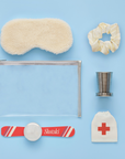 This image showcases the contents of the "Après Ski Set" laid out on a light blue background. The items include:
A soft, cream-colored sherpa sleep mask.
A satin scrunchie in a champagne color.
A collapsible metal shot glass.
A small pouch with a red cross symbol, likely for a hangover kit.
A mini "Shotski" accessory with a red, white, and blue stripe design.
A transparent pouch with a silver zipper to store all the items.