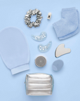 This image features a curated flat lay of self-care items arranged against a soft blue background. A blue exfoliating mitt,  A silver scrunchie, Glittery under-eye gel patches labeled "CHILL" and "OUT", A pair of cozy blue socks, A small white deodorant stick, A metallic gua sha facial tool, A silicone facial cleansing brush, A silver jar with an embossed star design (likely a skincare product) and a small metallic silver pouch.