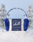 The image shows a winter-themed wellness kit with earmuffs prominently displayed. The earmuffs are plush with a shiny metallic band and bright blue fur. The packaging features the phrase "Permission to Chill" and mentions it as a "Winter Wellness Kit" by "Pinch Provisions." The background includes snowy elements and silver tinsel trees, creating a festive, wintry aesthetic.