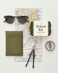 Flat lay image of travel essentials, including a pair of sunglasses, a green passport cover, a folding map, a compass, a pair of drafting dividers, and a green travel kit labeled '18 Essentials - Travel Kit for the Jet-Setter - Pinch Provisions.' The items are arranged on a light background for a clean and organized aesthetic.