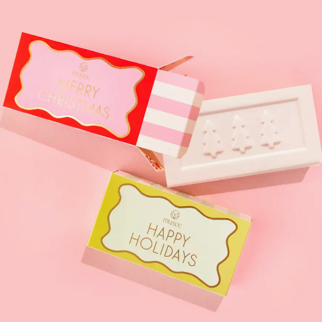 MUSEE MERRY CHRISTMAS SOAP WITH PINK AND WHITE STRIPED PACKAGING NEXT TO THE HAPPY HOLIDAYS SOAP