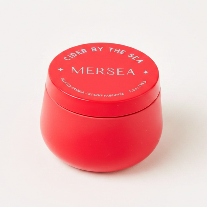 RED TIN CANDLE THAT READS, "CIDER BY THE SEA, MERSEA, SCENTED CANDLE" IN LIGHT BLUE WRITING