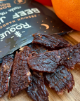 OPEN PACKAGE OF BEEF JERKY WITH AN ORANGE NEXT TO IT DISPLAYED ON A WOOD TABLE